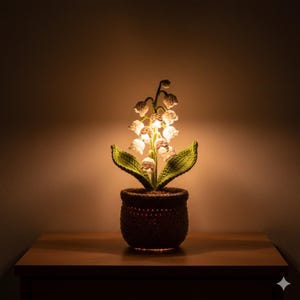 Könnte beinhalten: Gehäkelte Maiglöckchen-Lampe. Die Lampe hat weiße Blüten, grüne Blätter und einen braunen Topf. Die Lampe ist beleuchtet und strahlt ein warmes Licht aus. Die Lampe steht auf einer Holzoberfläche.