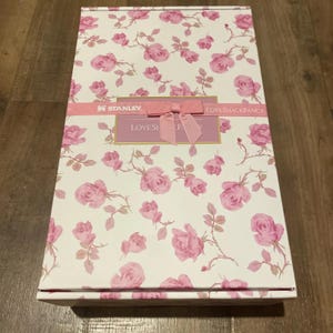 K&ouml;nnte beinhalten: Eine rechteckige Geschenkbox mit wei&szlig;em Hintergrund, bedeckt mit rosa Rosenmustern. Ein rosa Band mit einer Schleife umschlie&szlig;t die Box, mit dem Text "LOVESHACKFANCY" und "STANLEY". Die Box ist geschlossen.