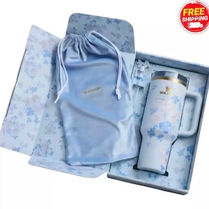 May include: A white Stanley mug with blue floral patterns, a handle, and a gold rim, presented in a floral-patterned gift box with a matching drawstring bag. The mug has the Stanley logo.