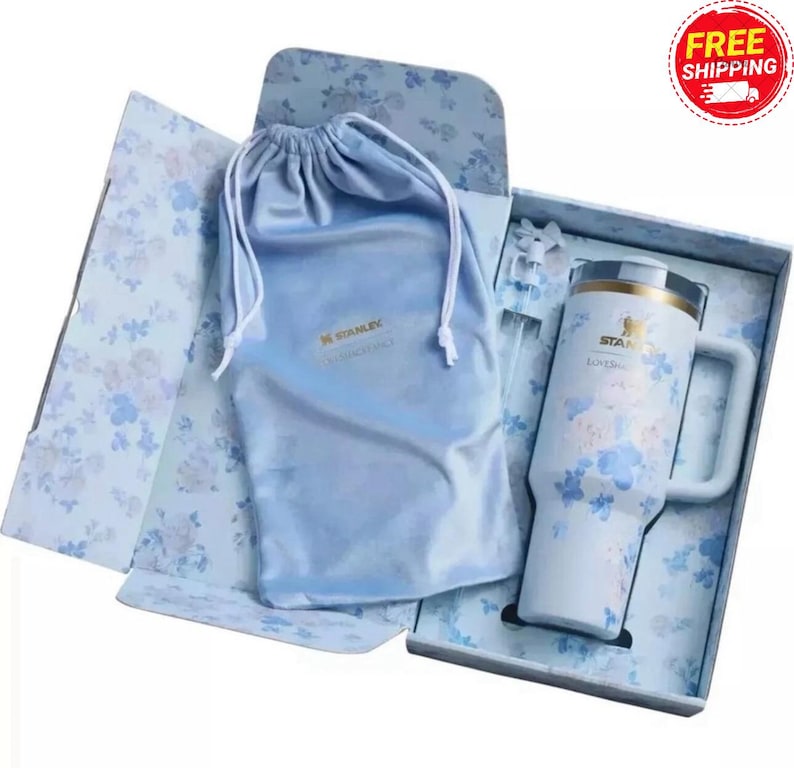 May include: A white Stanley mug with blue floral patterns, a handle, and a gold rim, presented in a floral-patterned gift box with a matching drawstring bag. The mug has the Stanley logo.