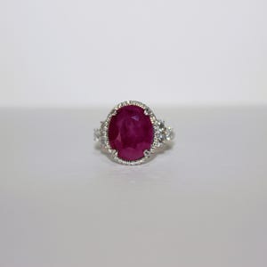 May include: A silver ring featuring a large, oval-cut ruby gemstone. The ruby is a deep, vibrant red color and is surrounded by a halo of smaller, clear gemstones. Additional gemstones are set on the ring's shoulders.