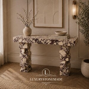 May include: A marble console table with a white and burgundy pattern. The table has a rectangular top and two stacked block legs. A vase with white flowers, a bowl, and books are on the table. The text "LUXURYSTONEMADE" is at the bottom.