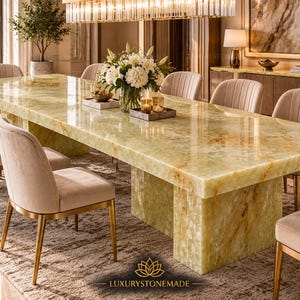 Puede incluir: Una gran mesa de comedor rectangular hecha de piedra de ónix verde claro pulida con vetas naturales, apoyada sobre una base de pedestal gruesa. La mesa está rodeada por ocho sillas beige tapizadas con patas de metal dorado y un logo abajo.