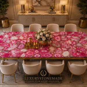 May include: A large, rectangular dining table with a vibrant pink agate stone top. The table is surrounded by eight cream-colored chairs with gold legs. A gold tray with candles and a vase of white flowers adorn the table. The table is from Luxury Stone & Epoxy Collection.