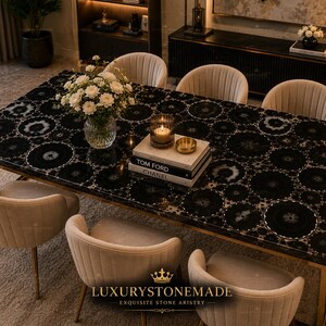 May include: A rectangular black dining table with a patterned surface, surrounded by six beige upholstered chairs. The table has a gold-colored metal frame. A vase of white flowers, candles, and books are on the table. The logo "LUXURYSTONEMADE" is at the bottom.