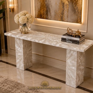 May include: A rectangular console table featuring a white, iridescent mother-of-pearl inlay arranged in a sunburst pattern across the top and legs. The table surface holds a glass vase with white flowers and a stack of three books labeled CHANEL, Dior, and TOM FORD.
