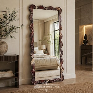 May include: A full-length mirror with a decorative, wavy frame. The frame has a dark red and white marble effect. The mirror reflects a bedroom with a bed, window, and neutral decor. A stylish home decor item.