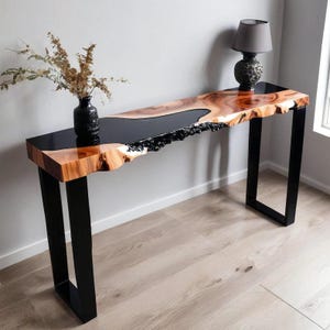 Puede incluir: Una consola de borde vivo con una incrustación de resina epoxi negra. La mesa presenta una tapa de vetas de madera natural y patas de metal negro. Un jarrón negro con flores secas y una lámpara decorativa están sobre la mesa. La mesa mide aproximadamente 152 cm de largo.
