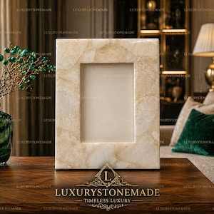 May include: A rectangular, off-white stone picture frame with a central opening. The frame is set on a dark wooden table, with a green vase and decorative branches to the left. The text "LUXURYSTONEMADE" is at the bottom.