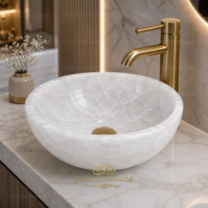 Puede incluir: Un lavabo de piedra blanca de forma redonda con una superficie cristalina texturizada y un grifo de metal dorado. El lavabo presenta un interior liso y un tapón de desagüe a juego en tono dorado, sobre una encimera de mármol de color claro.