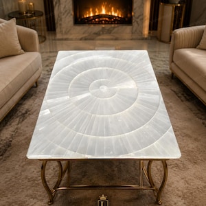 Op de afbeelding: Een rechthoekige salontafel met een blad van witte selenietsteen met een radiaal zonnestraalpatroon van ingelegde segmenten. De tafel wordt ondersteund door een goudkleurig metalen onderstel met gebogen poten en een horizontale steunbalk.