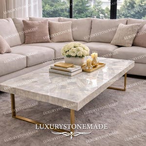 May include: A modern living room featuring a large, light-colored sectional sofa with decorative pillows. A rectangular coffee table with a white, patterned surface and gold-colored legs sits in front of the sofa. The text "LUXURYSTONEMADE" is visible.