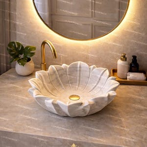 K&ouml;nnte beinhalten: Ein marmorner, bl&uuml;tenblattf&ouml;rmiger Waschbecken mit einer goldenen Armatur. Das Waschbecken steht auf einer Steinarbeitsplatte, mit einem runden Spiegel und einer kleinen Pflanze im Hintergrund. Der Text "LUXURYSTONEMADE" ist sichtbar.