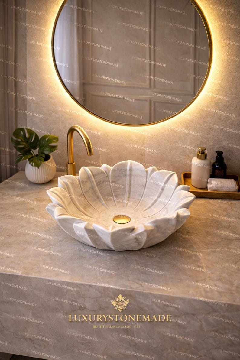 K&ouml;nnte beinhalten: Ein marmorner, bl&uuml;tenblattf&ouml;rmiger Waschbecken mit einer goldenen Armatur. Das Waschbecken steht auf einer Steinarbeitsplatte, mit einem runden Spiegel und einer kleinen Pflanze im Hintergrund. Der Text "LUXURYSTONEMADE" ist sichtbar.