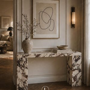 Puede incluir: Una consola de mármol con un patrón marrón y blanco. La mesa tiene una parte superior rectangular y dos patas cuadradas. Un jarrón con ramas, libros y un cuenco están sobre la mesa. Una obra de arte abstracta enmarcada cuelga de la pared encima de la mesa.