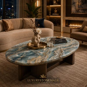 Op de afbeelding: Ovale salontafel met een blauw en goud marmeren blad en een donkere houten basis. De tafel staat in een woonkamer met een beige bank, kussens en een vaas met bloemen. De tekst "LUXURYSTONEMADE" staat onderaan.