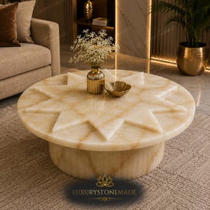 May include: A round coffee table crafted from light beige stone with a prominent star-shaped relief pattern on the surface. The table features a cylindrical base and a smooth, polished finish. A gold vase with white flowers sits on top.
