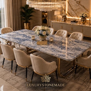 Puede incluir: Una mesa de comedor rectangular con tablero de mármol azul y blanco y base de metal dorado, rodeada por ocho sillas tapizadas en beige con detalles dorados. Un centro de mesa floral adorna la mesa, que incluye el texto LUXURYSTONEMADE.