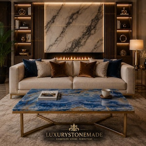 Op de afbeelding: Een luxe woonkamer met een crèmekleurige bank en fluwelen kussens in marineblauw, bruin en beige patronen. Een blauwe onyx salontafel met gouden poten staat voor de bank. De kamer heeft een open haard en inbouwkasten. De tekst "LUXURYSTONEMADE" is zichtbaar.