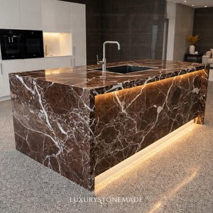 May include: A modern kitchen island made of brown marble with white veining. The island features a built-in sink and faucet, with a warm light strip along the base. The floor is a speckled gray, and the cabinets are white.