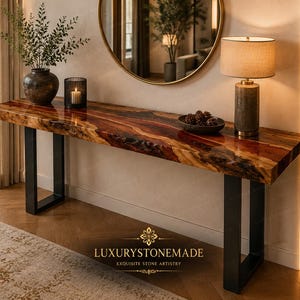 May include: A live edge console table with a reddish-brown wood grain finish. The table has black metal legs and is decorated with a lamp, a vase with greenery, and a candle. The table is part of the Luxury Stone Made collection.