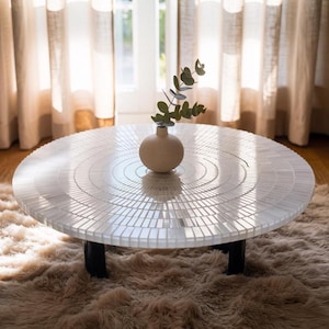 Puede incluir: Una mesa de centro redonda con una superficie de mosaico de azulejos blancos y patas negras. La superficie de la mesa presenta un diseño circular. Un pequeño jarrón beige con vegetación se encuentra sobre la mesa, colocada sobre una alfombra peluda.