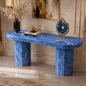 Puede incluir: Una mesa consola ovalada hecha de piedra azul vibrante con un acabado pulido, sostenida por dos pedestales hexagonales. El tablero presenta un patrón veteado natural y sostiene un jarrón negro con flores blancas, un adorno dorado y un libro.