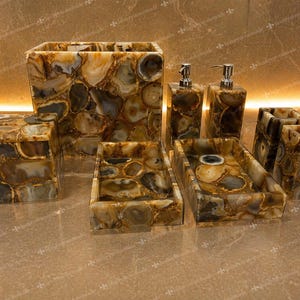 Puede incluir: Un conjunto de accesorios de baño hechos de piedra pulida y estampada. El juego incluye un dispensador de jabón, porta cepillos de dientes, un cesto de basura y bandejas. La piedra presenta una mezcla de tonos marrones, beige y grises, creando un aspecto natural y elegante.