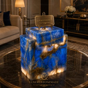 May include: A blue and gold stone lamp with a layered design. The lamp has a rectangular shape and is illuminated from within, creating a glowing effect. A small gold sign on top reads "LUXURYSTONEMADE".
