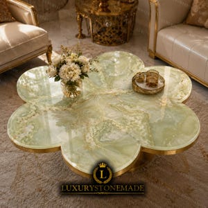 May include: A flower-shaped coffee table featuring a polished light green onyx stone top with natural white and gold veining, encased in a thin gold metal rim. A glass vase with white flowers and a small gold decorative tray sit on the surface.