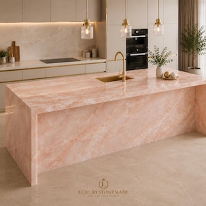 Puede incluir: Una cocina moderna con una gran isla de mármol rosa. La isla cuenta con un fregadero empotrado y un grifo dorado. Encima, hay lámparas colgantes con pantallas de vidrio. La cocina también incluye gabinetes blancos y un horno empotrado.