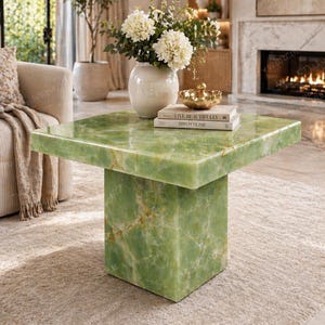 May include: A square, green onyx coffee table with a solid base. The table is topped with a vase of white flowers, a gold bowl, and two books. The table is in a living room setting.