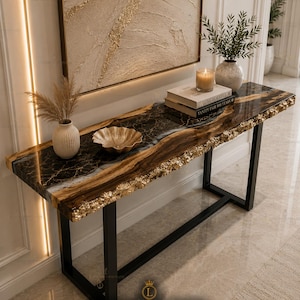 May include: A contemporary console table with a black metal frame and a distinctive tabletop. The tabletop has a black and gold epoxy resin design with a natural edge. Decorative items include a gold bowl, vases, books, and a candle. The wall art above the table has a gold textured design.