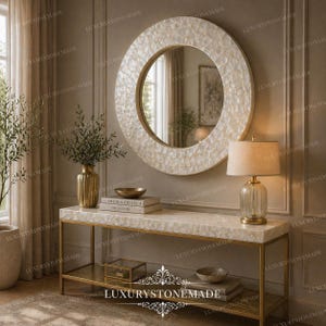 May include: A round mirror with a white mosaic-style frame hangs above a console table. The table has a gold metal frame and a white mosaic top. A gold lamp and vase are on the table, with books and a bowl. The room has a neutral color palette.