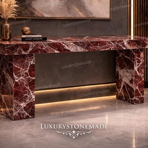 May include: A rectangular red marble console table with white veining. The table has two square legs and a matching top. A gold vase with dried flowers, a small gold sphere, and books are on the table.