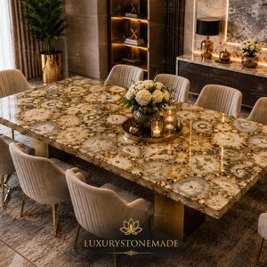 Puede incluir: Una mesa de comedor rectangular con una superficie pulida hecha de numerosas rodajas circulares de piedra ágata translúcida en tonos marrón y beige. La mesa se apoya sobre una base dorada y está rodeada por ocho sillas tapizadas en beige.