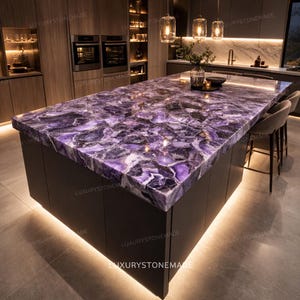 Può includere: Un'isola cucina con un ampio piano di lavoro in pietra preziosa viola lucidata. Il piano di lavoro presenta un motivo naturale unico con sfumature di viola, bianco e nero. L'isola ha mobili scuri ed è illuminata da luci a soffitto.