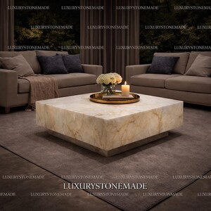 May include: A square marble coffee table with a light beige and gold veined pattern. A gold tray with flowers and a lit candle sits on top. The table is in a living room setting.
