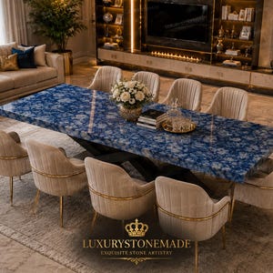 May include: A large, rectangular dining table with a blue and white patterned surface, surrounded by eight beige upholstered chairs with gold accents. The table is set with a floral arrangement and decorative items. The room features a neutral color palette and elegant decor.