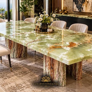 May include: A rectangular dining table featuring a polished green onyx stone top with white and brown veining, supported by two thick, cylindrical stone pedestals with a textured, multi-coloured finish. Includes a logo reading LUXURYSTONEMADE.