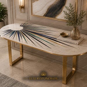 May include: An oval console table with a white marble top featuring a sunburst inlay pattern in blue, green, and black stone. The table has a gold-toned metal frame with two U-shaped legs. A book titled LIVE BEAUTIFULLY sits on the surface.