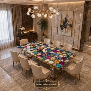 Könnte beinhalten: Ein luxuriöses Esszimmer-Set mit einem großen rechteckigen Tisch mit einer bunten Achatsteinplatte. Der Tisch ist von acht cremefarbenen Stühlen mit goldenen Akzenten umgeben. Über dem Tisch hängt ein goldener Kronleuchter.