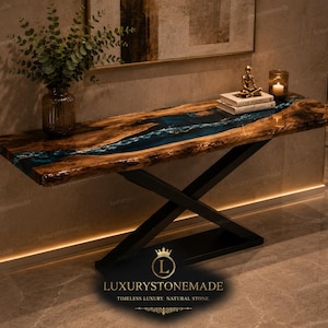 May include: A console table featuring a live-edge wood slab with a central blue resin river inlay, supported by a black metal X-shaped base. The table surface holds a glass vase with greenery, two stacked books, a small figurine, and a candle.