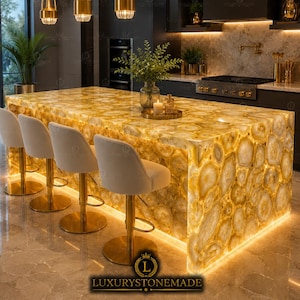 May include: A kitchen island featuring a backlit yellow agate stone countertop and base, paired with four white upholstered bar stools with gold-toned metal pedestals. The island surface displays a natural circular pattern of translucent stone.
