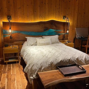 Può includere: Una camera da letto con una testiera in legno con un design a fiume in resina epossidica blu. La testiera è illuminata da un'illuminazione calda. Il letto ha un piumino e cuscini bianchi. Un comodino e una panca in legno sono in primo piano.