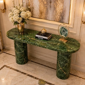 Op de afbeelding: Een lange, ovale consoletafel van groen marmer met gouden aders, ondersteund door twee cilindrische marmeren zuilen. Het oppervlak bevat een gouden vaas met witte bloemen, twee gestapelde boeken met de tekst Chanel en Tom Ford, en een agaat.