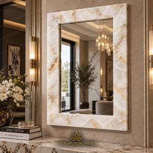 May include: A rectangular wall mirror featuring a polished frame with a white and cream marble pattern accented by gold veining. The mirror reflects an interior space with a chandelier and a potted plant, mounted on a matching marble wall.