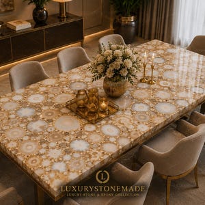 May include: A large rectangular dining table with a patterned surface of beige and white circular designs. The table is surrounded by beige upholstered chairs. A gold tray with decorative items and a vase of flowers are on the table.