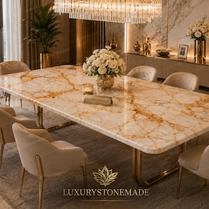 Op de afbeelding: Een rechthoekige eettafel met een gepolijst crèmekleurig en goud geaderd marmeren blad en een goudkleurig metalen onderstel. De tafel is omringd door acht beige gestoffeerde stoelen met gouden poten. Een bloemstuk draagt de tekst LUXURYSTONEMADE.