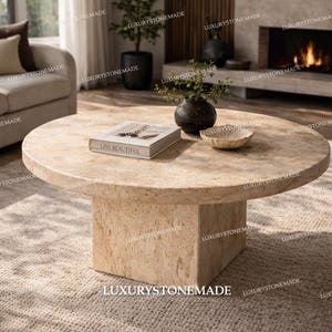 May include: A round, beige stone coffee table with a square base. The table has a book that reads "LIVE BEAUTIFUL", a small decorative bowl, and a dark vase with greenery. The table is on a neutral-colored rug.